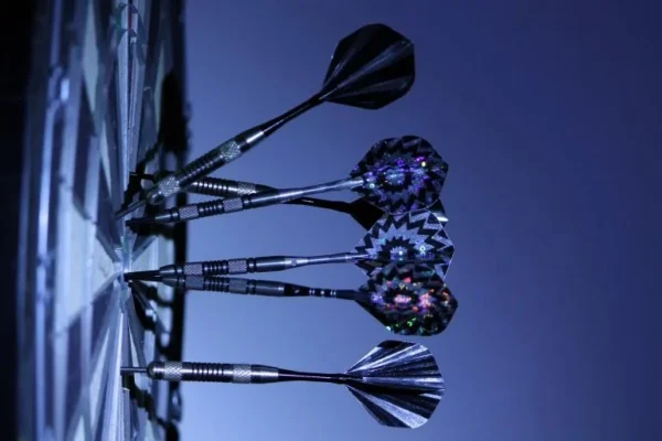 Vier Darts mit bunten Spitzen stecken in einer Dartscheibe, die in einem dunklen Raum aufgenommen wurde. Zielgenaues SEO
